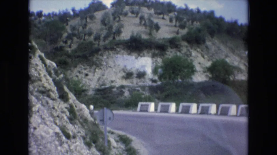 1972 Spain 8mm frame from a moving road view, showing a signpost at the curve, white concrete barriers, exposed hillside rock, and scattered olive-like trees.