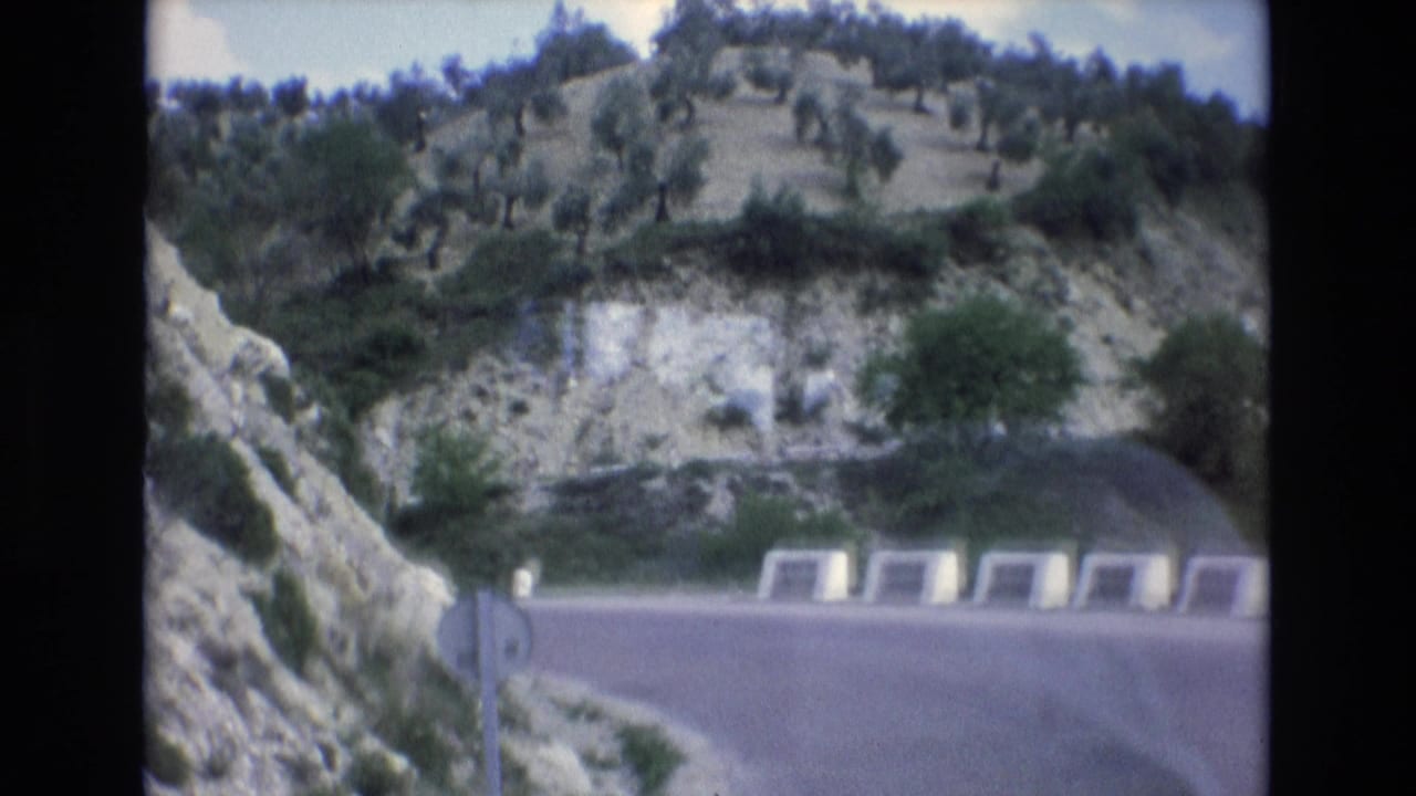 Winding road beside rocky hills with white roadside barriers and a circular sign.