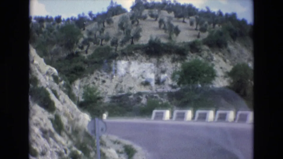 1972 Spain 8mm travel footage showing a curving asphalt road through rocky hills, with pale roadside barriers, shrubs, hillside trees, and a circular road sign.