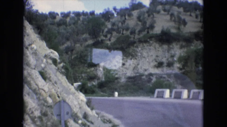 1972 Spain home-movie frame of a road bend bordered by white concrete barriers, a rocky slope, shrubs, and scattered trees on the hillside.