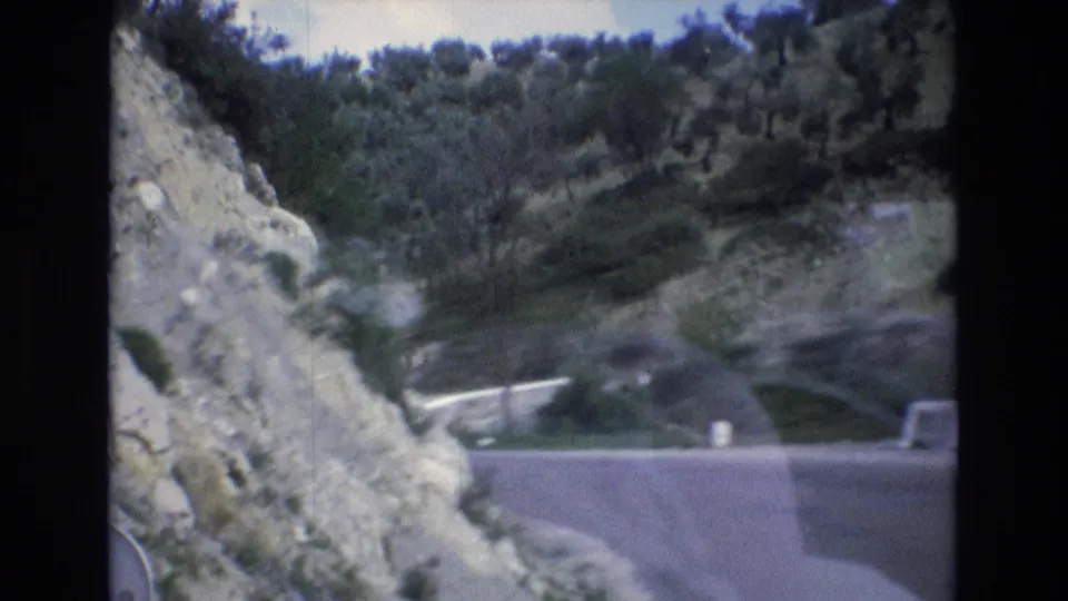 1972 Spain 8mm road-trip view with a rocky roadside close to the camera, a paved road, pale barriers, shrubs, and a green hillside in the distance.