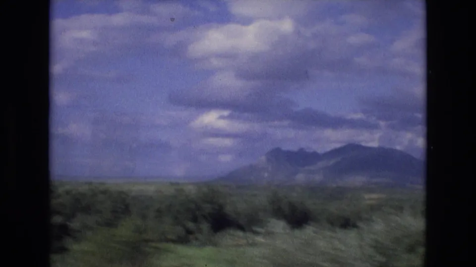 1972 Spain travel frame showing open countryside with low green vegetation, distant mountain ridges, and a clouded sky, likely filmed from a moving vehicle.