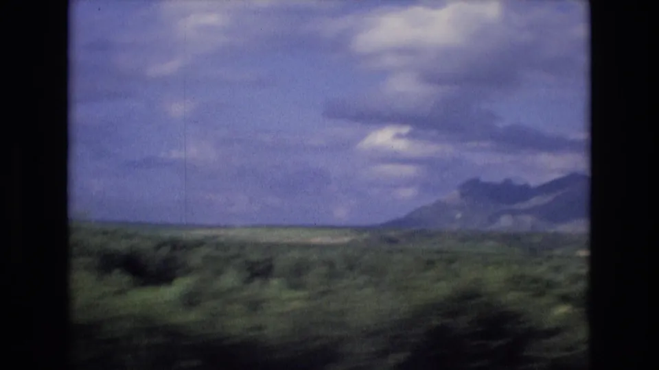1972 Spain home-movie frame of open countryside, low shrubs, and mountains under a dramatic cloudy sky, filmed from a moving vehicle.