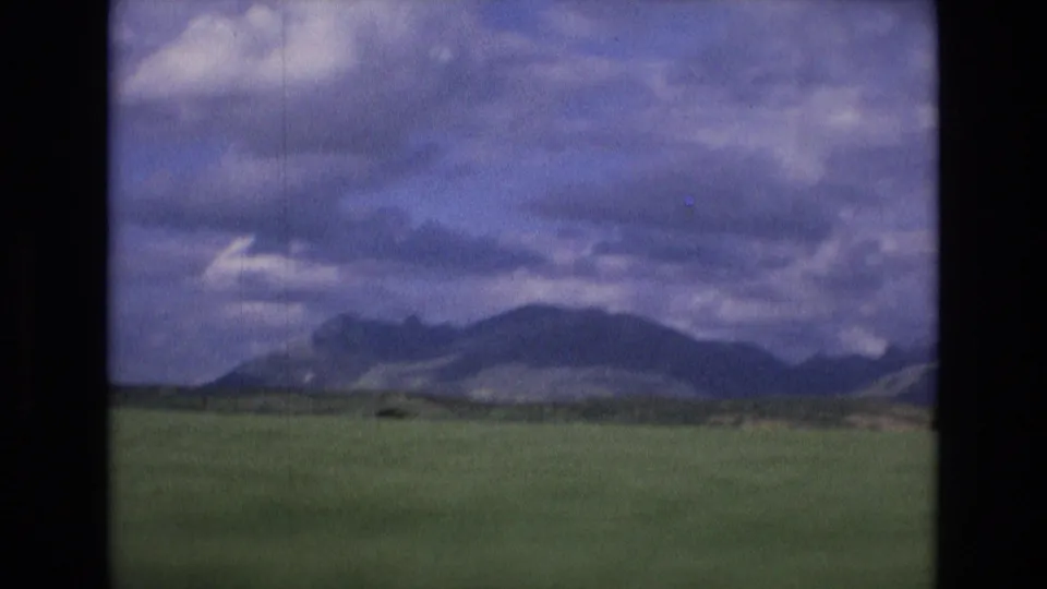 1972 Spain 8mm landscape view with a wide green field, distant mountains, and layered clouds, recorded from a moving vehicle.