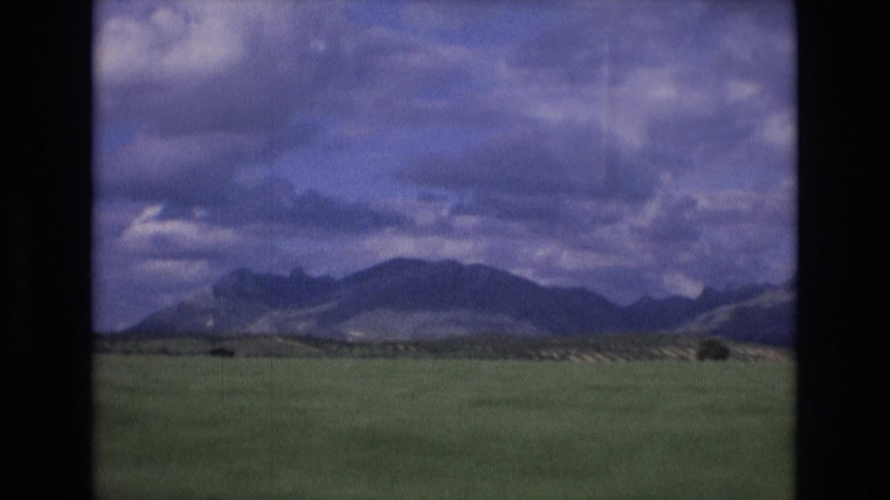 1972 Spain 8mm frame showing a broad green field stretching toward distant mountains under a cloudy blue-purple sky.
