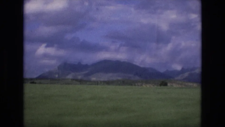1972 Spain 8mm travel view of an open field and low mountain range, framed by dark film edges and a mottled cloudy sky.