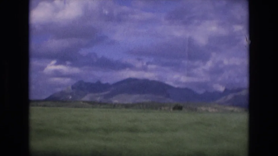 1972 Spain 8mm frame with green countryside in the foreground, distant hills along the horizon, and a textured cloudy sky above.