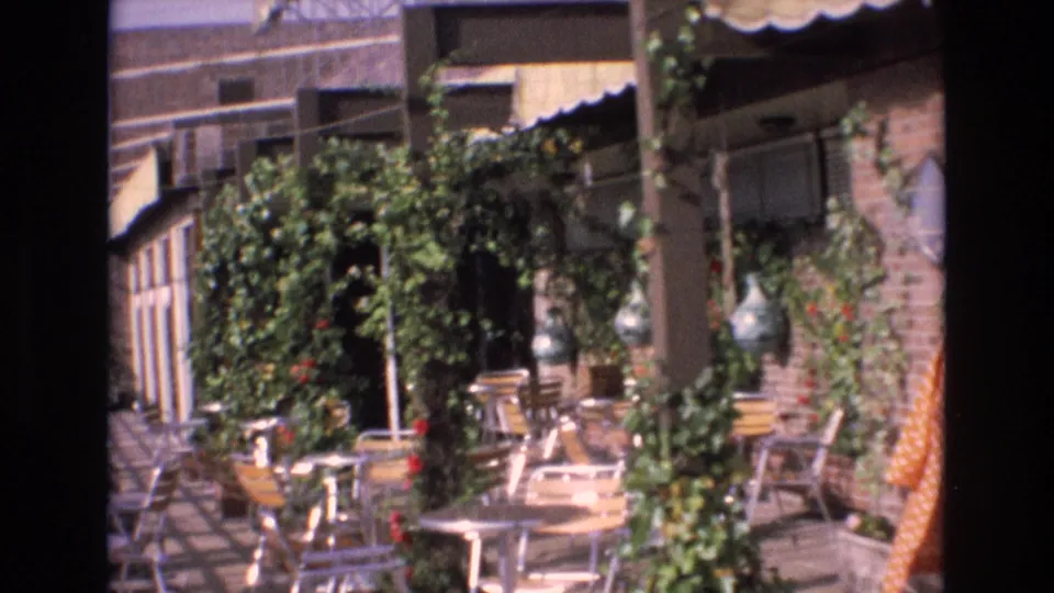 1972 Spain 8mm frame showing a vine-covered exterior seating area with tables, chairs, awnings, and brick or stone building surfaces.