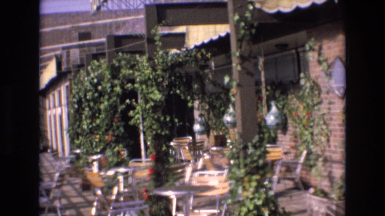 Spain 1972 8mm frame showing an empty restaurant patio with metal tables and chairs beneath green vines and a striped awning.