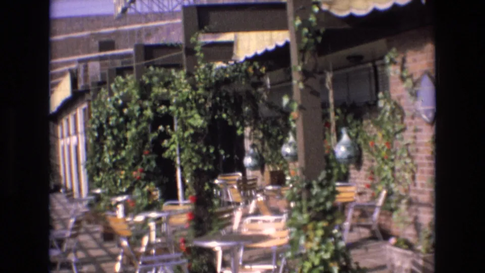 Spain 1972 8mm frame showing an empty restaurant patio with metal tables and chairs beneath green vines and a striped awning.