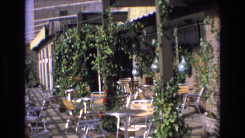 Spain 1972 home-movie frame of a sunlit outdoor dining area with empty metal furniture, leafy trellis growth, and decorative hanging glass bottles.