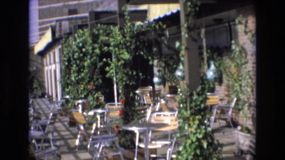 Spain 1972 8mm still showing empty café-style patio furniture surrounded by dense greenery, brickwork, and a shaded awning.