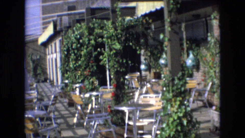 Spain 1972 travel-film frame of an empty outdoor restaurant patio with round metal tables, chairs, climbing vines, brick walls, and a striped awning.