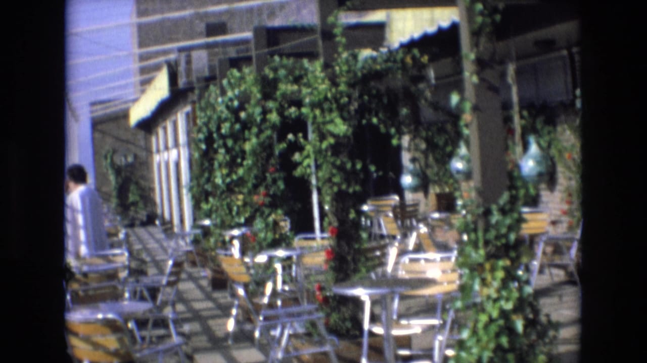 1972 Spain 8mm frame showing a sunlit rooftop cafe patio with metal tables and chairs, climbing greenery, yellow awnings, and a person partly visible at the left edge.