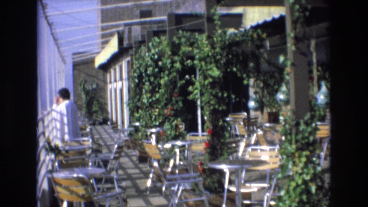 Outdoor terrace with empty chairs, round tables, dense greenery, and a person near the left side.