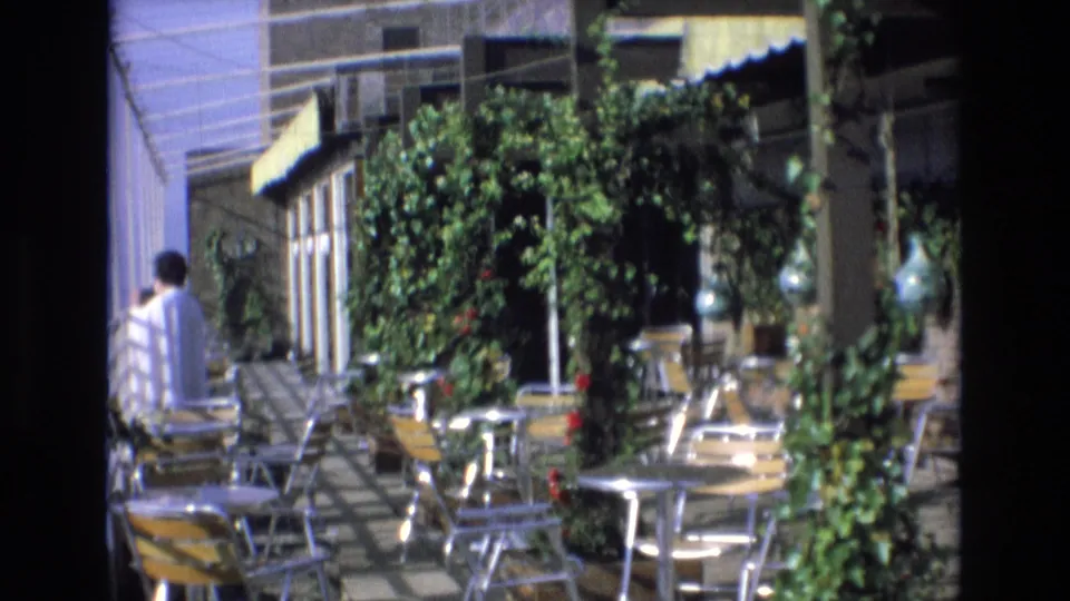 1972 Spain 8mm frame showing cafe patio seating arranged on tiled paving, with leafy trellises, potted plants, yellow awnings, and a person partly cropped at left.