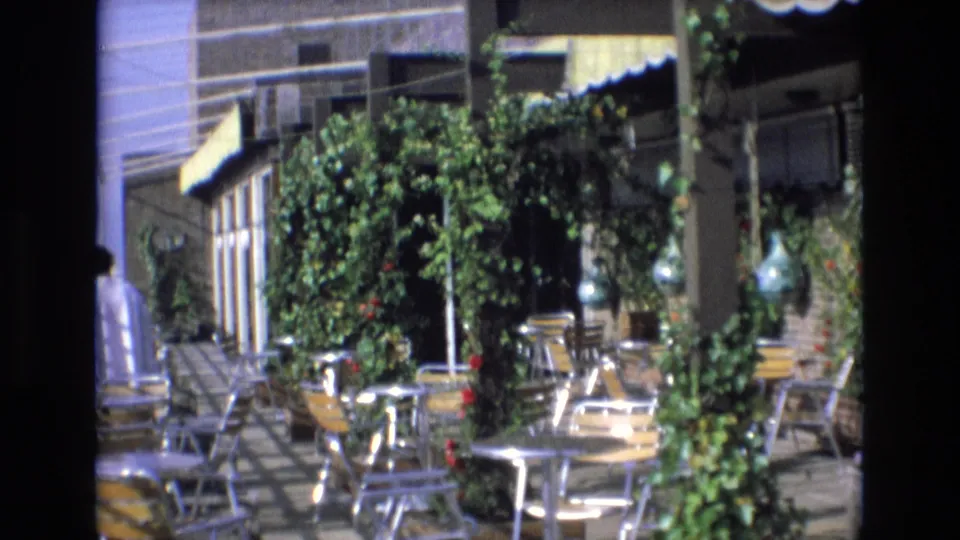 1972 Spain 8mm frame of a sunlit rooftop cafe terrace with metal tables, chairs, vines, potted greenery, yellow awnings, and building windows in the background.