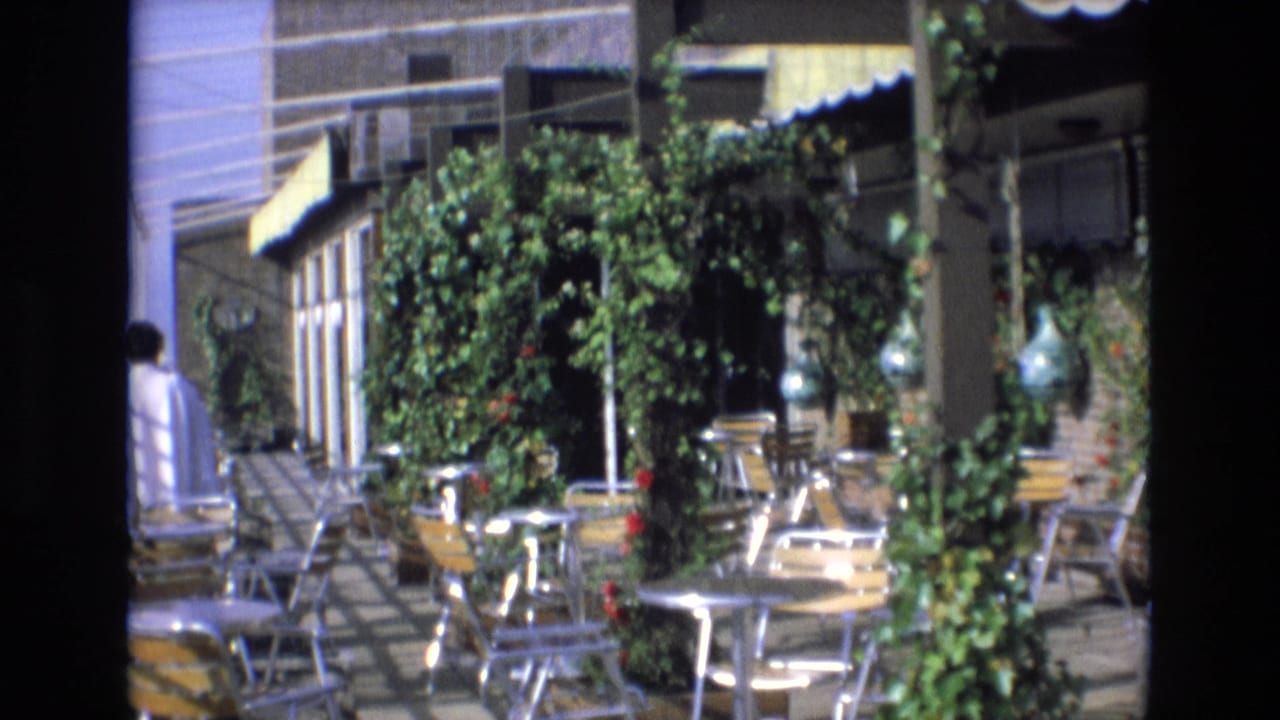 1972 Spain 8mm frame showing a sunny outdoor restaurant patio with metal chairs, round tables, climbing greenery, striped awnings, and a person partly visible at the left edge.