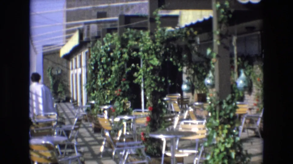1972 Spain 8mm frame of an outdoor cafe terrace with metal seating, round tables, dense vines, awnings, and a partial figure near the left side.