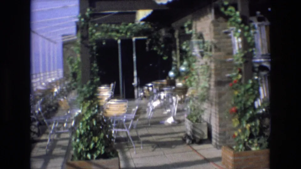 1972 Spain 8mm frame showing a patio restaurant area with stacked metal chairs, round tables, a brick pillar, climbing plants, planters, and dark doorway or window openings.