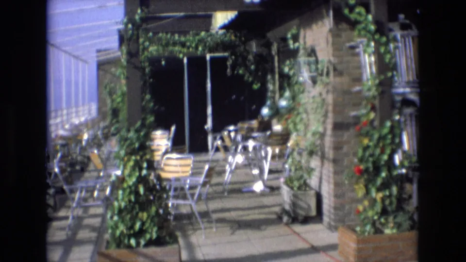 1972 Spain 8mm frame of an outdoor restaurant patio with metal cafe chairs, round tables, brickwork, climbing vines, potted greenery, and striped awnings in bright sun.
