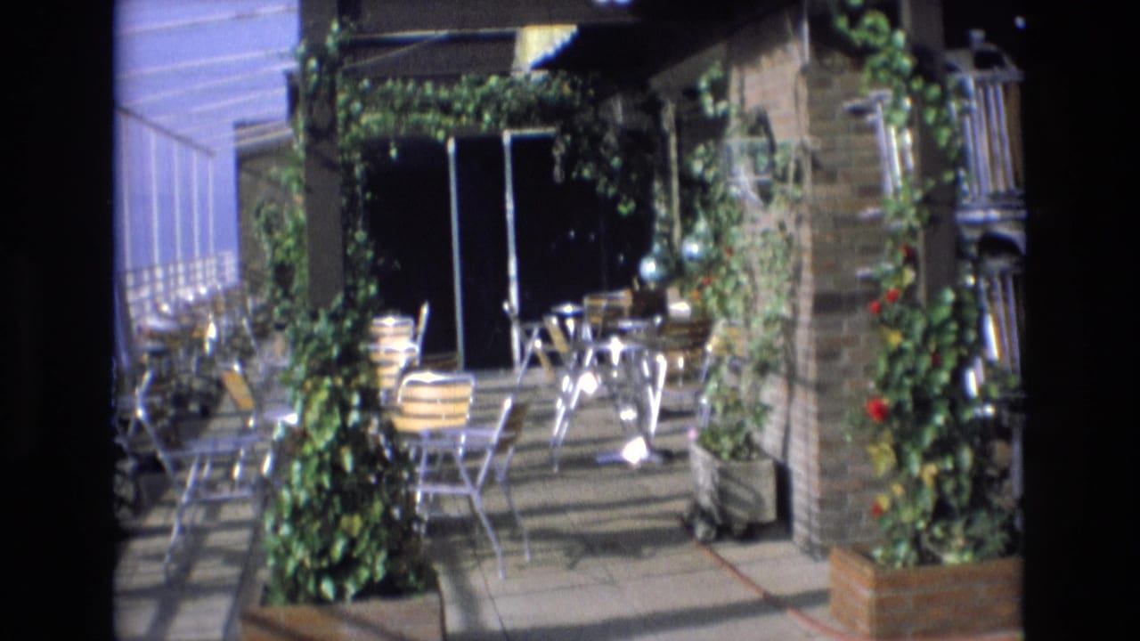 1972 Spain 8mm frame showing a rooftop or terrace patio with brick columns, climbing greenery, potted plants, striped chairs, and small tables.