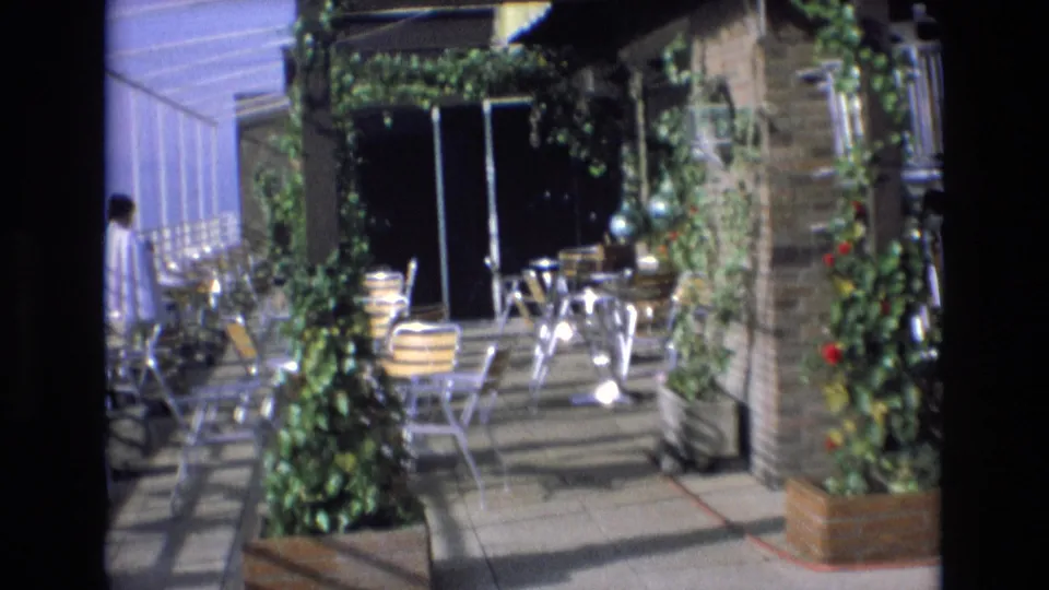 1972 Spain 8mm frame of a planted rooftop terrace with metal chairs, tables, brickwork, and a person partly visible at the edge of the scene.