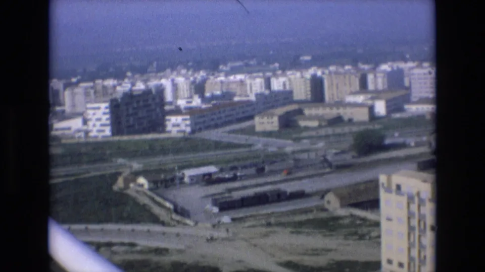 1972 Spain 8mm frame showing an elevated city view with dense apartment blocks, rail lines, roads, open ground, and industrial-looking buildings beneath a hazy sky.