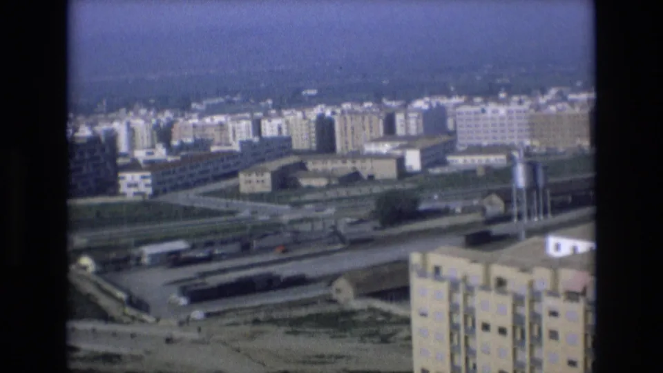 1972 Spain 8mm frame with a wide elevated view over apartment buildings, transport corridors, rail-yard structures, and hazy urban outskirts.