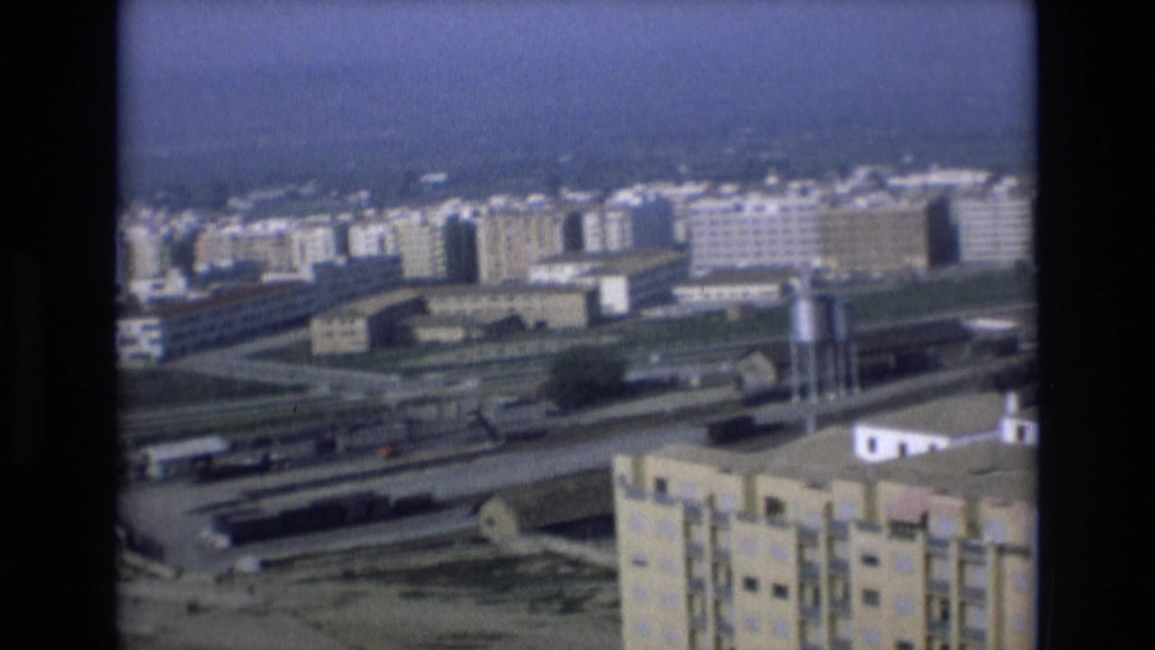 Elevated 1972 Spain city view with concrete apartment blocks, rail lines, rooftops, and a small utility tower seen through soft 8mm film grain.