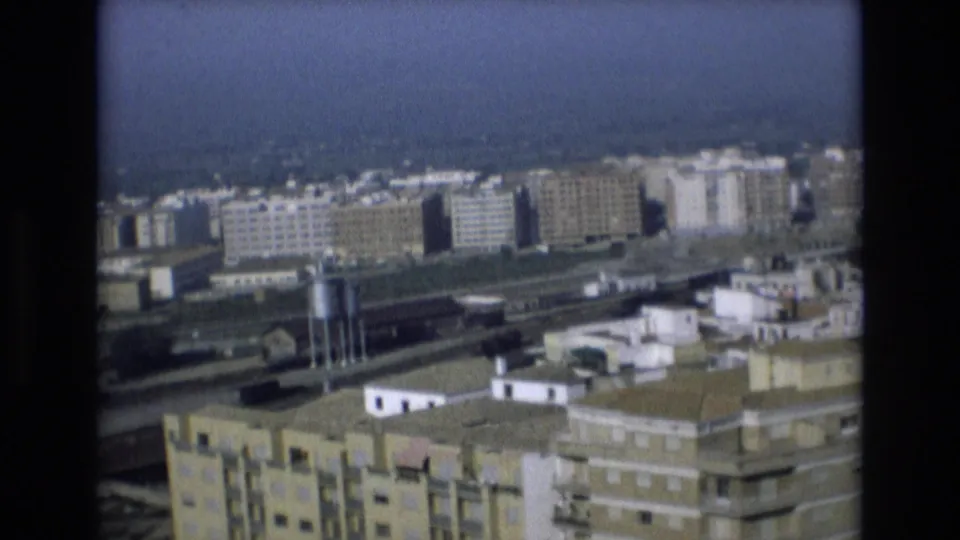 Elevated 1972 Spain cityscape with railway lines, blocky concrete housing, low rooftops, and a blue-gray horizon captured on soft 8mm film.