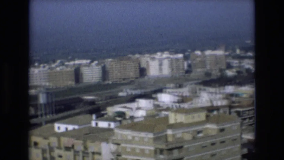 Late-frame 1972 Spain elevated view with concrete rooftops in the foreground, apartment blocks beyond, and rail infrastructure cutting through the cityscape.