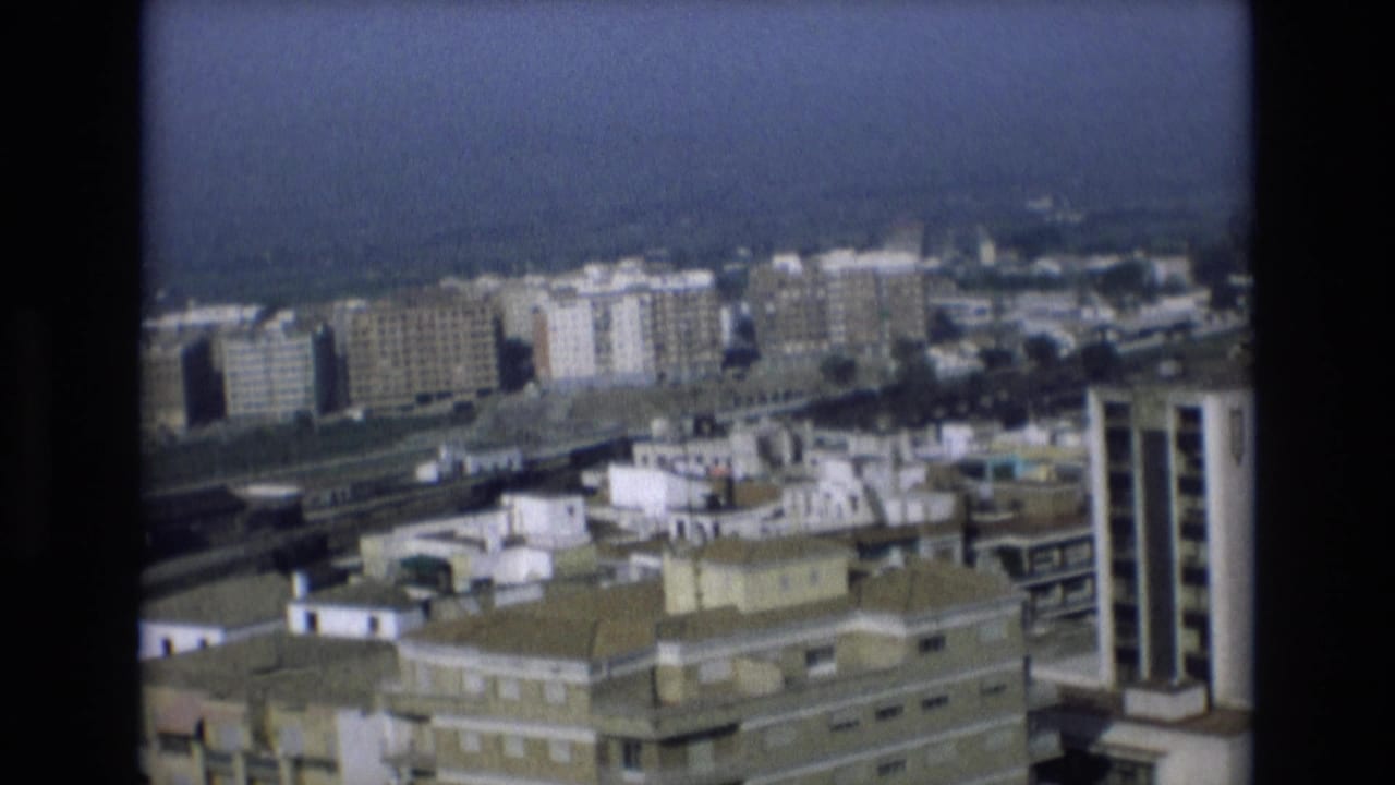 1972 Spain home-movie frame showing a hazy coastal cityscape with mid-rise apartment blocks, flat rooftops, and sea visible beyond the buildings.