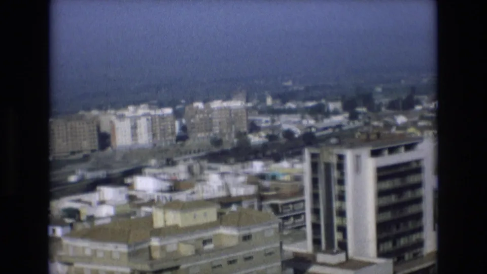 1972 Spain frame from a high viewpoint, with crowded city rooftops, a prominent white high-rise, and a hazy waterfront in the distance.