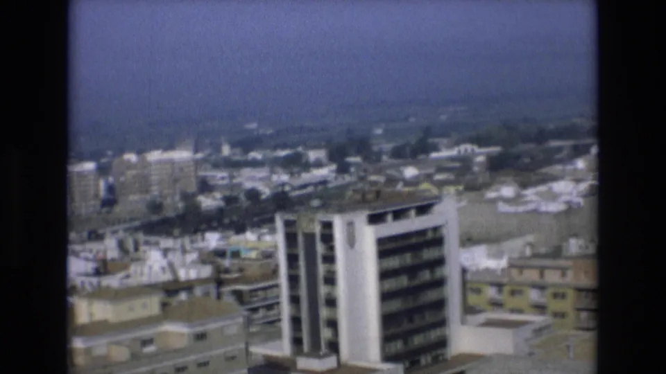 1972 Spain home-movie view across dense apartment buildings and flat roofs toward a soft blue sea horizon.