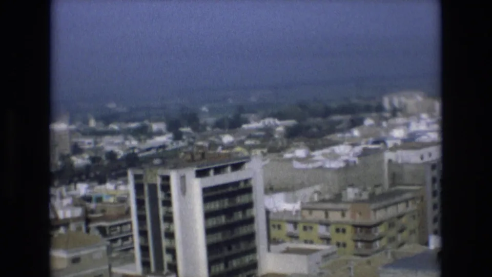 Final 1972 Spain frame showing a white high-rise, lower apartment blocks, rooftops, and the hazy coastline beyond the city.