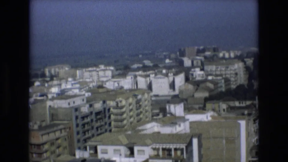 Final 1972 Spain cityscape frame showing clustered apartment buildings, flat roofs, balcony rows, and a muted blue-gray sky from an elevated angle.
