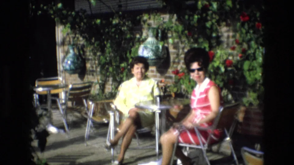 Two adult women in colorful 1970s dresses sit at a small patio table in Spain, surrounded by greenery, red flowers, and a brick wall.