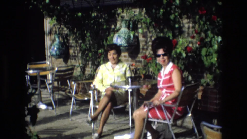 Two adult women on a sunlit Spanish patio look toward the home-movie camera, with metal chairs, drinks or tableware, plants, and a brick wall behind them.