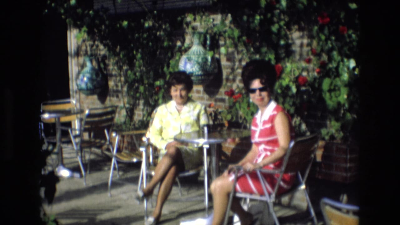 In Spain in 1972, two middle-aged women sit at a small metal outdoor cafe table, with one in a yellow dress and the other in a red-and-white striped dress and sunglasses.