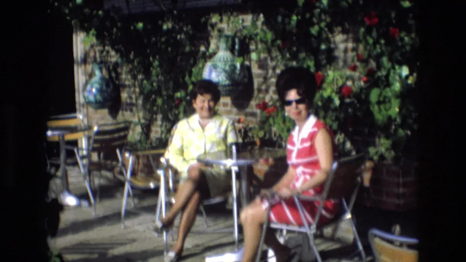 In the supplied 1972 Spain footage, the women remain seated at the outdoor cafe, framed by rose bushes, pottery vases, and garden furniture in bright sunlight.