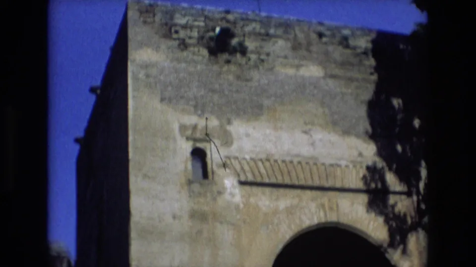 1972 Spain 8mm travel frame tilted upward on the stone structure, showing the upper wall, small arched opening, decorative slats, blue sky, and dark side shadows.