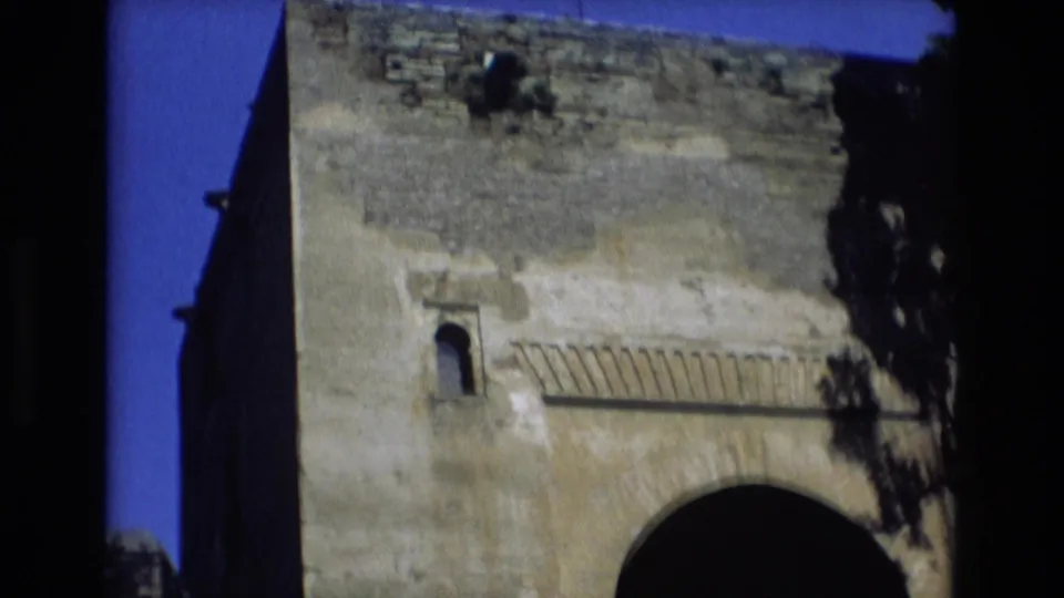 1972 Spain 8mm frame showing the upper facade of a weathered stone structure, with a small arched window, horizontal decorative detail, and the top of a large dark arch.