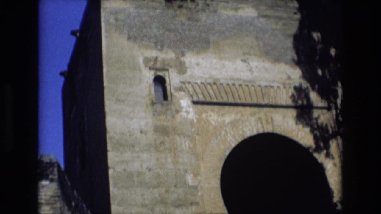 1972 Spain 8mm frame showing a pale historic stone wall with a dark arched opening, small upper window, and blue sky at the edge.