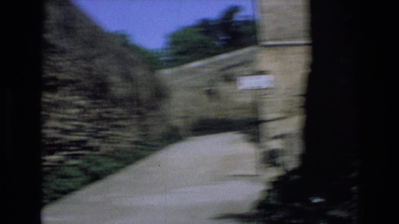 1972 Spain home-movie frame showing a blurred narrow road between high stone walls, seen from a dark doorway-like edge with sky and greenery beyond.