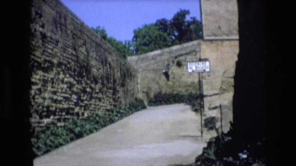 1972 Spain home-movie frame showing a sunlit narrow lane bordered by weathered stone walls, greenery, blue sky, and a visible street sign.