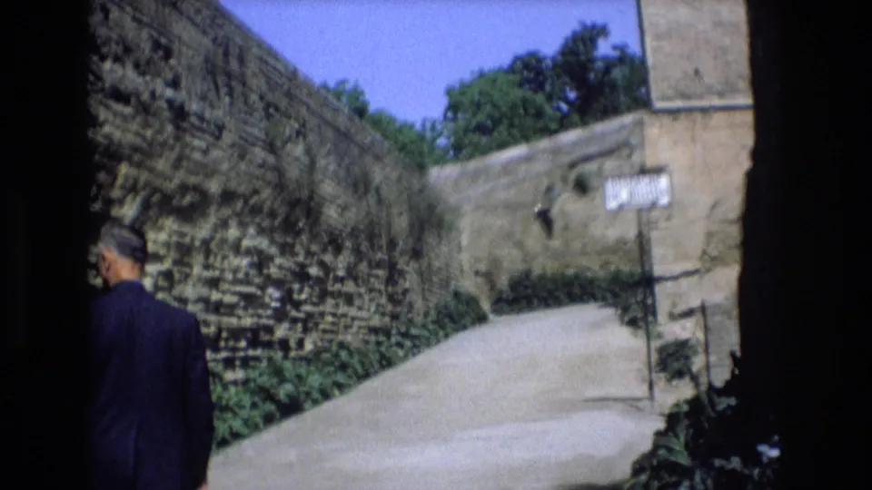 1972 Spain home-movie frame showing an adult man in dark clothing walking away beside a high weathered wall on a narrow sunlit lane.