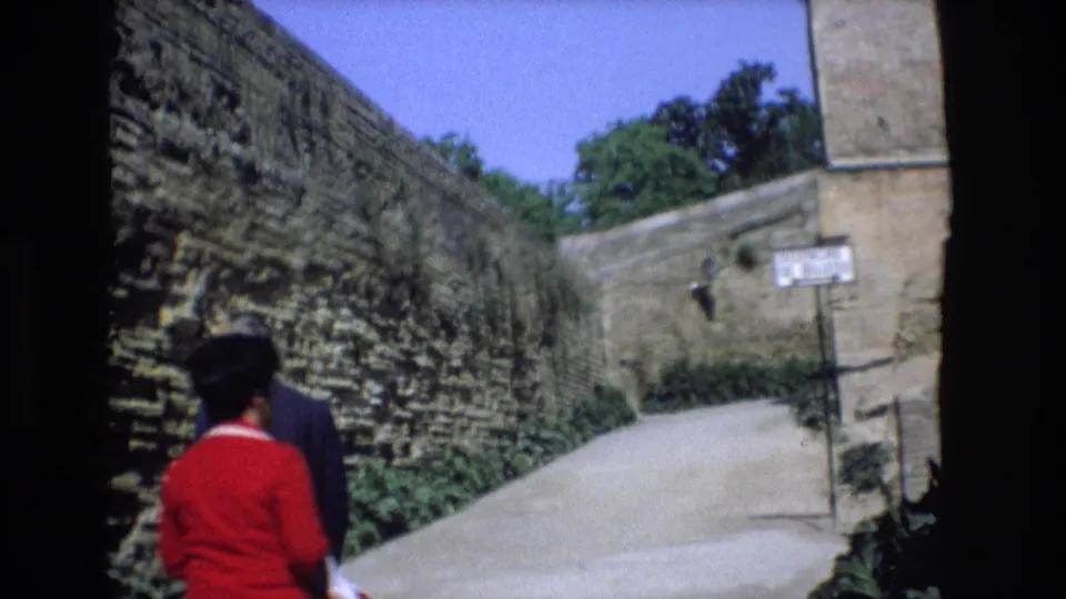 1972 Spain home-movie frame showing two adults, including a woman in red and a man in dark clothing, walking away along a narrow lane between stone walls.