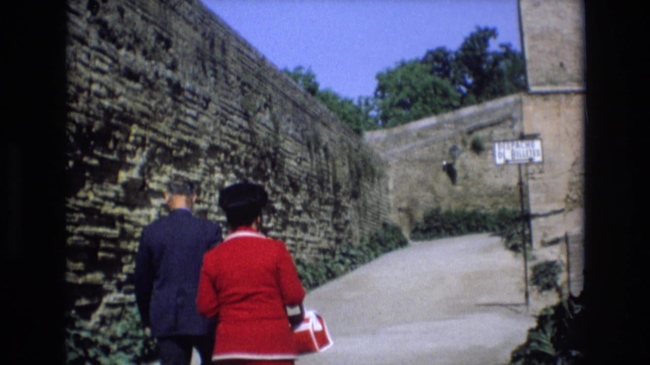 In 1972 Spain home-movie footage, adults walk away from the camera along a path bordered by high weathered stone walls and greenery.
