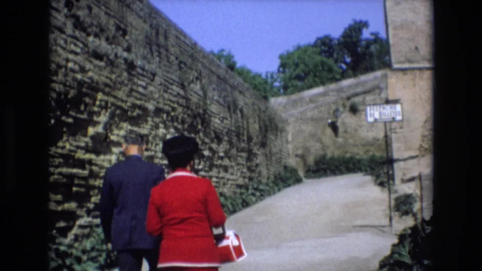 In 1972 Spain home-movie footage, adults walk away from the camera along a path bordered by high weathered stone walls and greenery.